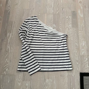 Randig one shoulder topp från Kappahl - Säljer en vit och marinblå randig topp från Kappahl i 100% bomull. Modellen är one shoulder med lång ärm på ena sidan och axelbandslös på andra, vilket ger en snygg asymmetrisk look. 