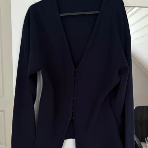 Shaped knitted top från Gina - Snygg mörkblå blus med lång ärm och djup v-ringning. Blusen har knappar framtill som en snygg detalj och är i ett mjukt, stickat material. Perfekt för dig som gillar stilrena och enkla plagg med en twist.