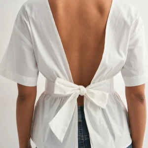 Vit blus med öppen rygg och rosett - Supersöt vit blus i bomull med korta ärmar, djup öppen rygg och stor knytrosett bak i midjan. Blusen har en markerad midja och lätt volang nedtill för en feminin siluett. Prislapp kvar!!