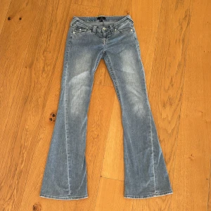 True Religion Joey Low Rise Flare Jeans - Säljer ett par blåa True Religion Joey jeans med låg midja och utsvängda ben. Klassisk femficksmodell med snygga kontrastsömmar och ikoniska bakfickor med lock och knapp. Jeansen är i denim och har en cool, lite sliten tvätt. Perfekta för dig som gillar retrovibbar!