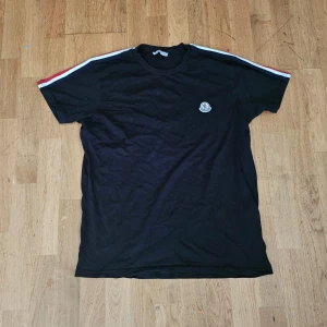 Svart Moncler t-shirt - Svart t-shirt från Moncler i bomull med korta ärmar. Snygga röda, vita och blå ränder längs axlarna och ett broderat Moncler-märke på bröstet. Klassisk rund halsringning och normal passform. Perfekt för dig som gillar stilrena plagg med en sportig touch.