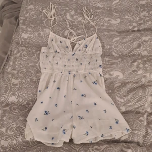 Vit blommig jumpsuit med knytband - Supersöt vit jumpsuit med tunna axelband som knyts och smockad överdel. Jumpsuiten har små blå blommor broderade över hela tyget och är gjord i ett lätt bomullstyg. Perfekt för varma dagar och ger en romantisk vibe. Har en liten ljus gul fläck men inget man ser! Kan sälja billigare🎀