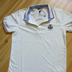 Vit Moncler pikétröja med blå krage - Vit pikétröja från Moncler med blå insida på kragen och röda detaljer på ärmsluten. Klassisk Moncler-logga broderad på bröstet. Skön passform och perfekt för en clean och sportig look. pris kan diskuteras