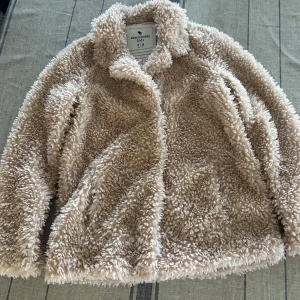 Beige teddyjacka Abercrombie Kids - Fluffig och supermjuk teddyjacka från Abercrombie Kids i beige färg. Jackan har klassisk krage, långärmad design och är tillverkad i syntetmaterial som ger en cool och mysig look. Perfekt för dig som gillar chill och trendiga vibes.