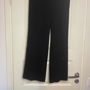 Svarta vida kostymbyxor från Wera - Snygga svarta kostymbyxor från Wera i storlek 38. Byxorna har en rak och vid passform med pressveck framtill för en clean look. Perfekta för dig som gillar stilrena och trendiga plagg. Materialet känns mjukt och följsamt.