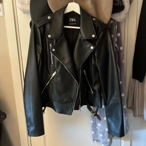 Svart skinnjacka från Zara - Snygg svart skinnjacka från Zara i storlek XL men passar mig som har ca M så liten i storleken. Perfekt nu till hösten! Klassisk bikerstil med silvriga dragkedjor, knappar och bälte i midjan. Jackan har flera fickor med dragkedja och detaljer som axelklaffar. Den har inte kommit till användning därför säljer jag den🖤