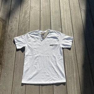 Vit Essentials Fear of God t-shirt - Vit t-shirt från Essentials Fear of God med svart logga tryckt på bröstet. Klassisk rund hals och korta ärmar. Tillverkad i mjuk bomull för en clean och stilren look. Perfekt för dig som gillar minimalistisk streetwear. Storlek S men ganska baggy pris kan diskuteras 
