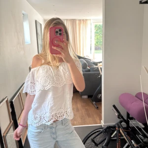 Vit offshoulder blus med broderi - Superfin vit offshoulder blus med broderade detaljer och hålmönster längs volangkanten. Luftig och lätt, perfekt för varma dagar. Blusen har en lös passform och är ärmlös med bred volang som täcker axlarna.