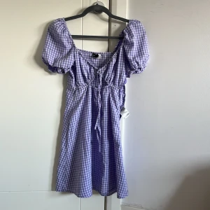 Lila rutig klänning med puffärm - Superfin lila och vit rutig klänning med puffiga korta ärmar och söt snörning framtill. Klänningen har en smickrande midja och lätt utställd kjol, perfekt för en somrig look. Tillverkad i mjukt bomullstyg som känns skönt mot huden.