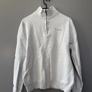 Ljusgrå stickad half zip från Bondelid - Snygg ljusgrå stickad tröja från Bondelid med half zip och hög krage. Broderad logga på bröstet och ribbade muddar vid ärmslut och nederkant. Perfekt för lager-på-lager och chill vardagsstil.