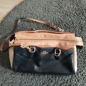 Coach axelväska i skinn, beige & svart - Snygg axelväska från Coach i en mix av beige och svart skinn med guldiga metalldetaljer. Väskan har justerbar axelrem, dragkedja upptill och rymligt fack inuti. Perfekt för dig som vill ha en klassisk men ändå trendig look.