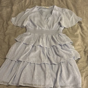 Ljusblå volangklänning från NA-KD - Supersöt ljusblå kortklänning från NA-KD med v-ringning, korta puffärmar och flera lager volanger. Klänningen har ett prickigt mönster och smock i midjan som ger en snygg siluett. Perfekt för dig som gillar romantisk och feminin stil.