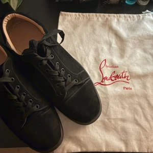 Louboutin  - Säljer ett par svarta sneakers från Christian Louboutin med snygg snörning och rund tå. Utsidan är i mjuk mocka och insidan har beige skinnfoder. Diskret broderad logga på hälen ger en lyxig vibe. Perfekt för dig som gillar stilrena och exklusiva skor. Har lite problem på kanterna