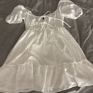 Vit kortklänning med puffärm Gina Tricot - Supersöt vit kortklänning från Gina Tricot i storlek S. Klänningen har puffärmar, snörning framtill och volang nedtill. Materialet är lätt och krinklat, vilket ger en chill och romantisk vibe. Perfekt för varma dagar och sommarhäng.