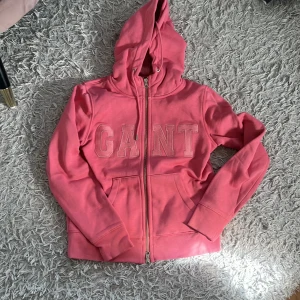 Rosa hoodie  - Säljer en snygg rosa hoodie från GANT med stor logga framtill. Tröjan har huva med snörning, dragkedja hela vägen och två fickor. Perfekt för chill dagar eller när du vill vara extra comfy. Materialet är mjukt och skönt, troligtvis bomullsmix.