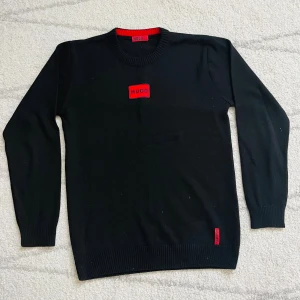 Svart tröja från Hugo med röd logga - Svart långärmad tröja från Hugo med röd logga på bröstet och nertill. Klassisk rund halsringning och ribbade muddar. Perfekt för dig som gillar stilren design med en touch av färg. Materialet känns mjukt och skönt mot huden.