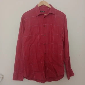 Röd rutig linneskjorta Rose & Born - Snygg röd skjorta från Rose & Born i storlek 40, tillverkad i linne. Skjortan har ett rutigt mönster med tunna vita linjer och klassisk krage. Långärmad modell med knappar framtill, perfekt för dig som gillar stilrena detaljer.