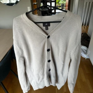 Beige kofta från H&M S - En stilren beige kofta från H&M i storlek S med svarta knappar framtill. Koftan har v-ringning, långa ärmar och är tillverkad i ett mjukt stickat material. Regular fit.