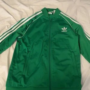 Grön Adidas track jacket med vita ränder - Säljer en grön Adidas track jacket med klassiska vita ränder längs ärmarna och Adidas-logga på bröstet. Jackan har dragkedja framtill, ståkrage och två fickor. Tillverkad i glansig polyester för en sportig vibe.