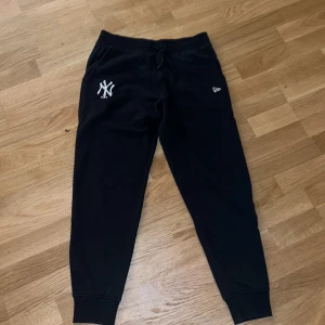 Svarta mjukisbyxor New Era Yankees - Svarta mjukisbyxor med New York Yankees-logga på vänster lår och New Era-märke på höger sida. Byxorna har snörning i midjan och muddar vid bensluten. Perfekta för chill eller sportig stil.