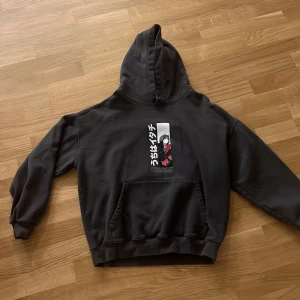 Svart Naruto Itachi hoodie - Svart hoodie med stor Itachi Uchiha-print från Naruto på både fram- och baksidan. Japansk text och Akatsuki-symboler i rött och vitt. Klassisk känguruficka och huva. Perfekt för animefans som vill sticka ut.