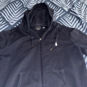 Mörkblå hoodie från Polo Ralph Lauren - Mörkblå hoodie från Polo Ralph Lauren med klassisk vit logga på bröstet. Tröjan har huva med snörning, hel dragkedja och två fickor framtill. Tillverkad i mjuk bomull, perfekt för chill dagar. Snygg och enkel design som funkar till allt.