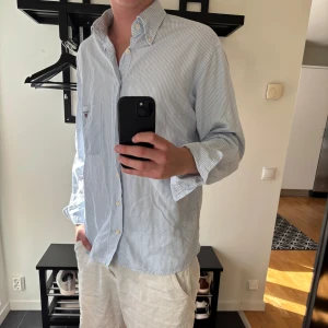Randig skjorta från GANT i bomull - Säljer en klassisk blå- och vitrandig skjorta från GANT i storlek L. Skjortan har button down-krage, bröstficka med broderad GANT-logga och är tillverkad i mjuk bomull. Perfekt till jeans eller chinos för en clean look.