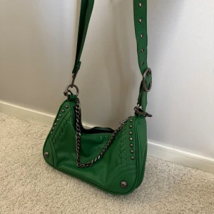 Grön axelväska med kedjedetalj från Zara - Cool grön axelväska i skinn med silverfärgade nitar och kedja. Väskan har justerbart axelband med stora ringar och dragkedja. Köpt från Zara, aldrig använd.