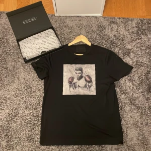 Svart limitato t-shirt med konstnärligt tryck - Svart t-shirt från limitato med ett stort konstnärligt tryck på bröstet som föreställer en abstrakt skalle i svart, vitt och rött. T-shirten har rund hals och korta ärmar, och är gjord i mjuk bomull för skön passform.