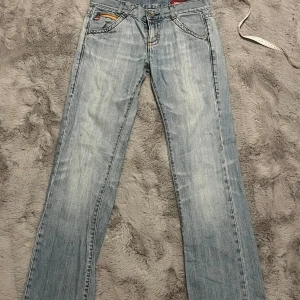 Miss Sixty ljusblå bootcut jeans - Snygga ljusblå jeans från Miss Sixty med klassisk femficksdesign, orange kontrastsömmar och en liten röd logga på bakfickan. Jeansen har bootcut-ben och låg midja, tillverkade i slitstarkt denim med dragkedja och knapp framtill. På baksidan så är en bältesögla lös samt små slitningar vid ena bakfickan(allt synd på bilderna) midjemått:37cm, grenhöjd:20cm, innerbenslängd: 78cm, torallängd: 100cm