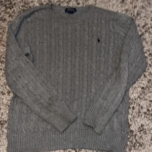 Grå stickad tröja från Polo Ralph Lauren - Kabelstickad grå tröja från Polo Ralph Lauren i storlek XL. Klassisk rund halsringning, ribbade muddar och diskret broderad logga på bröstet. Perfekt för dig som gillar stilrena och tidlösa plagg med premiumkänsla.