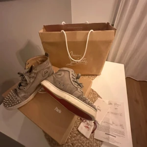 Christian Louboutin sneakers med nitar - Gråa high-top sneakers från Christian Louboutin i mocka med ikonisk röd sula och silverfärgade nitar på tån. Skorna har snörning och rund tå, samt diskret logga på sidan. Snygg och edgy design som sticker ut. Köpta för 740 euro och andvönda fåtal gånger 