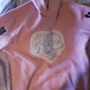 Rosa hoodie med tryck och huva - Säljer en rosa hoodie med stor vit och grå logga på bröstet och svart text längs båda ärmarna. Tröjan har huva och en stor magficka framtill. Perfekt för dig som gillar streetwear och vill ha en skön och snygg look.
