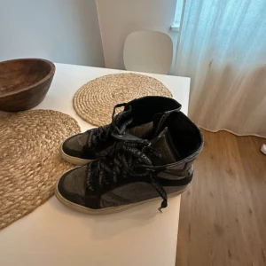 Svarta high top sneakers Zadig & Voltaire - Snygga high top sneakers från Zadig & Voltaire i svart och grått med glittrig finish. Skorna har snörning med logotyp, dragkedja på sidan och detaljer i skinn och glitter. Perfekta för dig som gillar en edgy stil. Några slitningar och en tärning men den märks inte av alls 