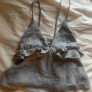 Randig linnetopp med volang och knyt - Superfin randig linnetopp med smala axelband, volangdetalj över bysten och knyt i ryggen. Toppen är vit och mörkblå, har en v-ringad form 