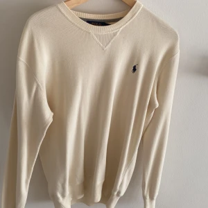 Beige tröja från Polo Ralph Lauren - Klassisk beige tröja från Polo Ralph Lauren med rund halsringning och broderad marinblå logga på bröstet. Tröjan är långärmad och tillverkad i mjuk bomull, perfekt för en clean och stilren look.