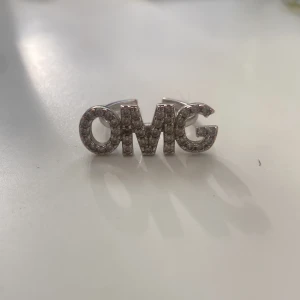 Silverfärgad OMG-ring - Cool statementring i silverfärg med texten 'OMG' framtill, täckt av små glittrande stenar. Ringen är öppen baktill och justerbar i storlek. Perfekt accessoar för dig som vill sticka ut och visa din personlighet. Den är från ioaku, nickelfri och rostar inte!