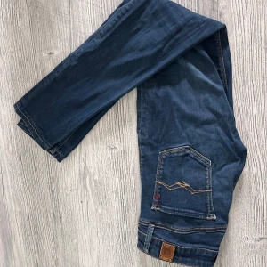 Replay Anbass jeans blå denim - Snygga Replay Anbass jeans i mörkblå denim med klassiska kontrastsömmar och cool broderad bakficka. Byxorna har normal passform och är tillverkade i stretchig bomullsblandning för extra komfort. Märkespatch i läder baktill och detaljer i rött vid fickan. Någon liten defekt 