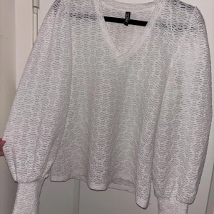 Vit spetsig blus från Pieces - Säljer en vit blus från Pieces med v-ringning och långärmade puffärmar. Blusen har ett genombrutet spetsmönster som ger en luftig och trendig känsla. Perfekt att styla med jeans eller kjol för en fräsch look.
