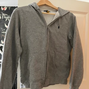Grå hoodie från Polo Ralph Lauren - Mysig grå hoodie från Polo Ralph Lauren med dragkedja och huva. Tröjan har mjukt teddyfoder på insidan, två fickor fram och klassisk broderad logga på bröstet. Perfekt för kyliga dagar. Storleken passar S/xs.