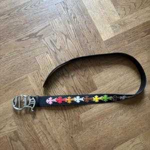 Chromehearts bälte - Coolt svart bälte i skinn med stora färgglada kors längs hela bandet. Spännet är i silverfärgad metall och har en unik design. Längd utan buckla 105 cm. Lite slitage vid hålen.