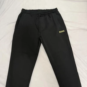 Svarta mjukisbyxor från BOSS - Svarta mjukisbyxor från BOSS med gul logga på vänster ben. Byxorna har elastisk midja med snörning och två sidofickor. Perfekta för en chill och avslappnad stil.