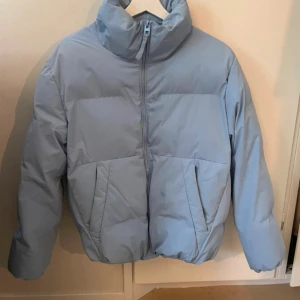Ljusblå pufferjacka med dragkedja - Säljer en ljusblå pufferjacka med hög krage och hel dragkedja framtill. Jackan har två stora fickor på sidorna och en oversized passform som ger en chill vibe. Perfekt för kalla dagar när du vill vara både varm och snygg.
