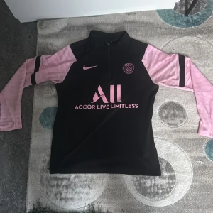 PSG långärmad Nike Dri-Fit tröja - Paris Saint-Germain fotbollströja från Nike med Dri-Fit material. Svart kropp med ljusrosa långärmade ärmar och rosa detaljer. PSG-logga och Nike Swoosh på bröstet, samt 'Accor Live Limitless' och 'Visit Rwanda' tryck. Halv dragkedja framtill och modern passform.
