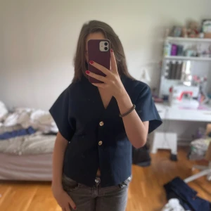 Mörkblå kortärmad blus med guldknappar - Pris kan duskuteras!! Säljer en stilren mörkblå blus med korta ärmar och två snygga guldknappar framtill. Toppen har en rak, boxig passform och är gjord i ett stickat material som känns både mjukt och lite tjockare. Den är insydd i midjan så den sitter taigthare där, alldrig använd. 