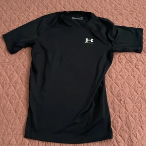 Svart kompressionst-shirt Under Armour - Helt ny oanvänd svart kompressionst-shirt från Under Armour med logga på bröstet och en etikett för namn på ryggen. Tight passform och tillverkad i stretchigt syntetmaterial, perfekt för träning. Klassisk rund hals och korta ärmar. Mindre i storleken! 