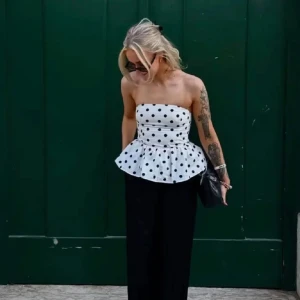 Vit prickig bandeau topp med volang - Supersöt axelbandslös bandeau topp i vit bomull med svarta prickar och volang nertill. Toppen har en figurnära passform upptill och en utställd peplumkant som ger en snygg siluett. Perfekt att styla med jeans eller kjol för en trendig look.