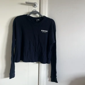 Mörkblå långärmad tröja Newport - Mörkblå ribbad långärmad tröja från Newport med vit text på bröstet och stor tryckt text på ryggen. Tröjan har en croppad passform och är tillverkad i mjukt bomullsmaterial. Perfekt för en avslappnad och sportig stil.