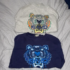 2 st Kenzo sweatshirt med tigertryck - Två Kenzo sweatshirts med ikoniskt broderat tigerhuvud på bröstet. En är marinblå med blått och vitt broderi, den andra är ljusgrå med färgglatt broderi i blått, orange, grönt och gult. Båda har rund hals och lång ärm, tillverkade i mjuk bomull.