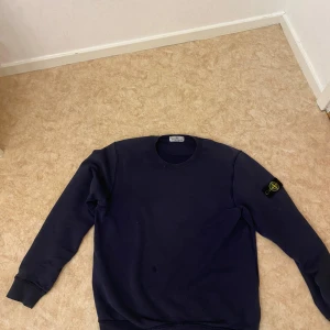 Mörkblå Stone Island sweatshirt - Mörkblå sweatshirt från Stone Island med klassisk rund hals och ikonisk patch på vänster ärm. Tröjan har ribbade muddar och är tillverkad i mjuk bomull för en skön och avslappnad stil. Perfekt för dig som gillar streetwear och snygga detaljer.